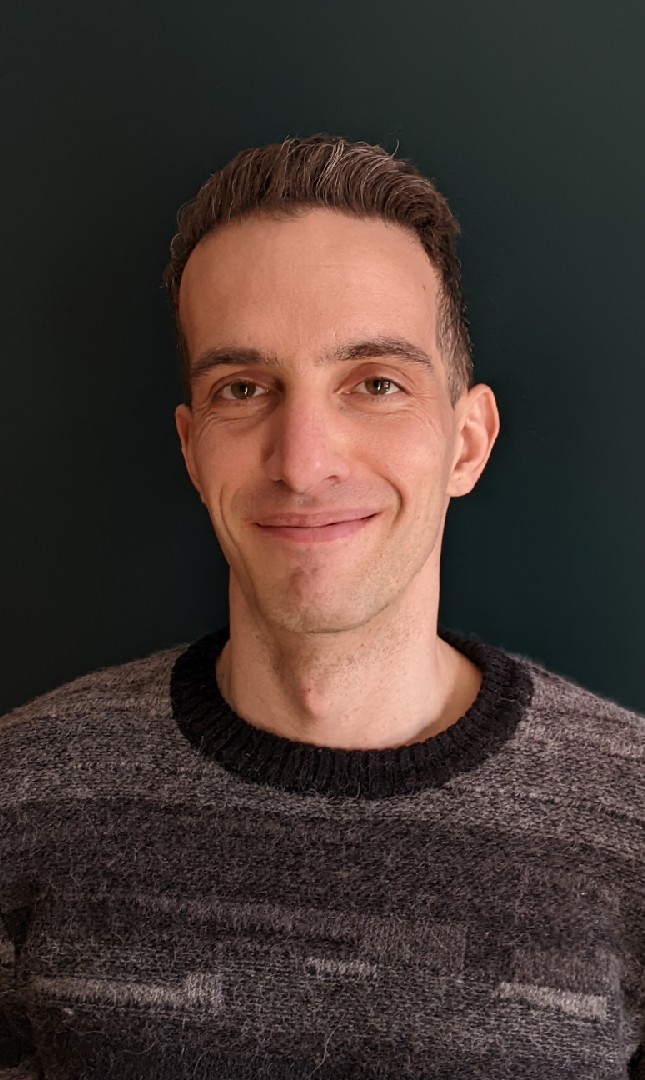 A caucasian man in his thirties, with wavy dark hair with some gray streaks wearing a dark gray sweater